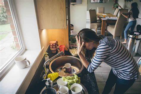 An overwhelmed mom leans over a sink full of dirty dishes, holding her head in her hand. Behind her, children run in circles around the dining table, adding to the chaos. Her posture and expression show pure exhaustion in the midst of a hectic household.